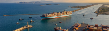 An aerial, wide-angle 16:9 view of a massive container ship cruising through a calm blue waterway towards an open sea under a soft, golden sunset sky. In the foreground, a bustling Mediterranean shipping port features stacks of multicolored cargo containers and smaller support vessels docked along the pier. A stone breakwater with a small lighthouse guides the ship, while distant hills line the horizon, capturing a strategic maritime gateway. Gemini return to Suez could strengthen Eastern Mediterranean gateways