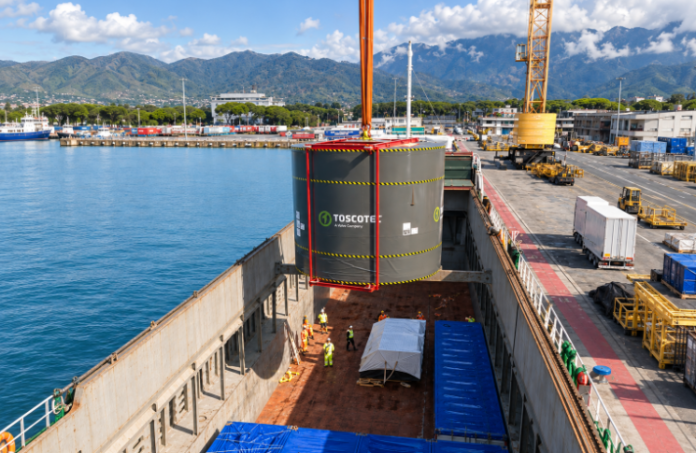 A port crane loads a large industrial cargo unit into a vessel’s open hold at a mountainous coastal terminal.