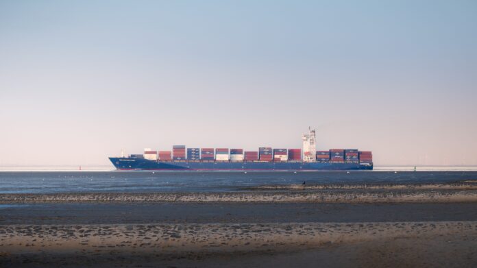 A container ship carrying stacked containers sails along a calm coastal waterway beneath a hazy sky.