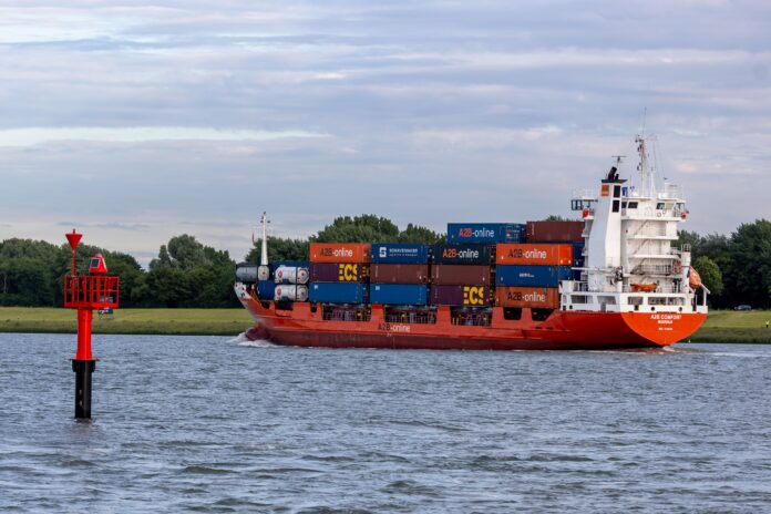 A2B Comfort container ship carrying stacked cargo containers sails along a waterway past a red navigation marker.